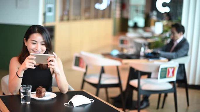 一名亚洲中国女性在自助餐厅茶歇期间享受甜点巧克力蛋糕，同时练习社交距离，用她的智能手机玩网络游戏