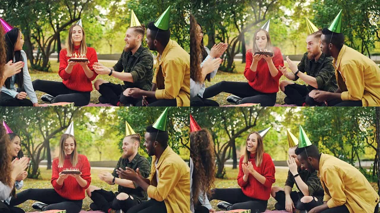 戴着派对帽的漂亮女孩正在公园野餐时与朋友一起庆祝生日，许愿，在蛋糕上吹蜡烛，开心地笑。事件和自然概念