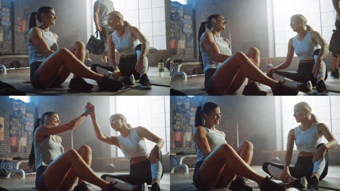两个美丽健康的运动女孩坐在工业阁楼健身房的地板上。他们对自己的培训计划感到满意，并成功地击掌。强壮的