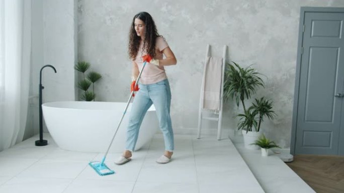 快乐的年轻女士用拖把洗地板，在浴室里在家跳舞