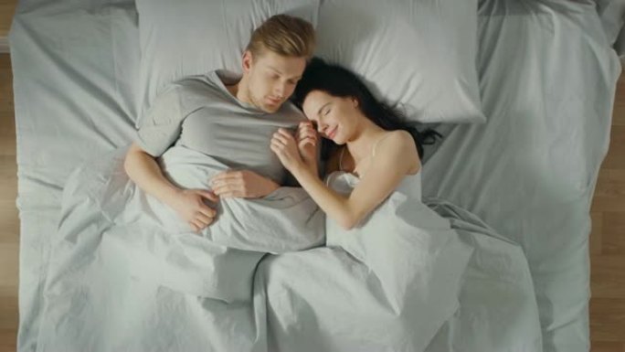 幸福的年轻夫妇在床上拥抱在一起。美丽的女孩和英俊的男孩在悠闲的早晨，慢慢地醒来并在早晨伸展。自上而下