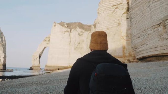 后视图高加索旅游男子背着背包沿着海边走到阳光明媚的埃特雷塔慢动作的白色拱形悬崖。