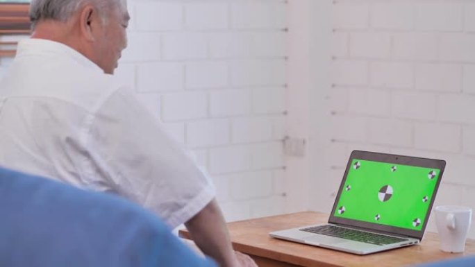 亚洲老爷爷在家用电脑看绿屏到视频会议。家里绿屏