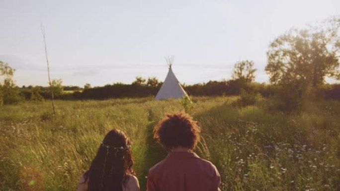 年轻夫妇在野营度假时穿过田野走向圆锥形帐篷的后视图 -- 慢动作拍摄