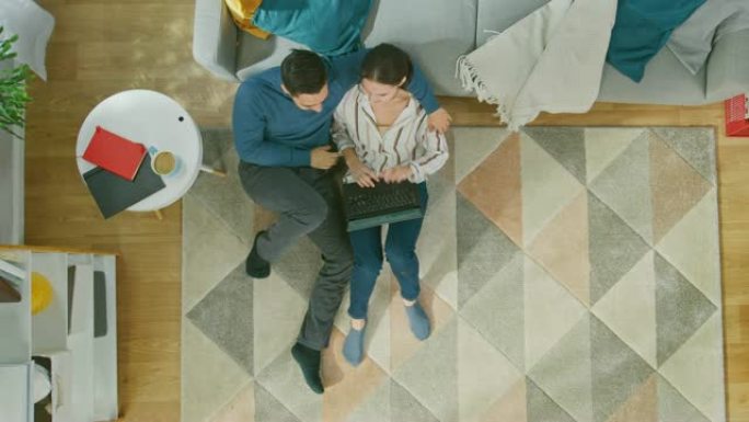 年轻夫妇坐在地板上，用笔记本电脑积极交谈。舒适的客厅，配有现代室内地毯、沙发、椅子、桌子、架子、植物