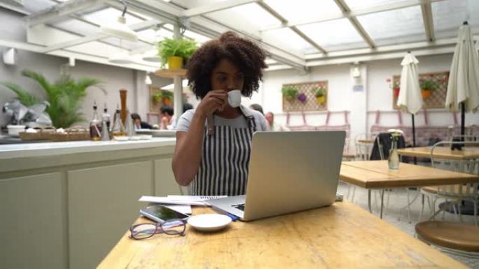 一家餐厅的美丽黑人企业家老板在笔记本电脑上工作时喝咖啡