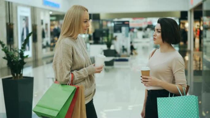 美丽的年轻女性在商场里聊天，拿着外卖饮料和纸袋，讨论购买和分享新闻。青年生活方式和商店概念。