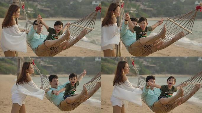 可爱的亚洲家庭一起在海滩吊床上享受。暑假期间，年轻的亚洲夫妇和小儿子一起躺在吊床上的慢动作镜头，日落