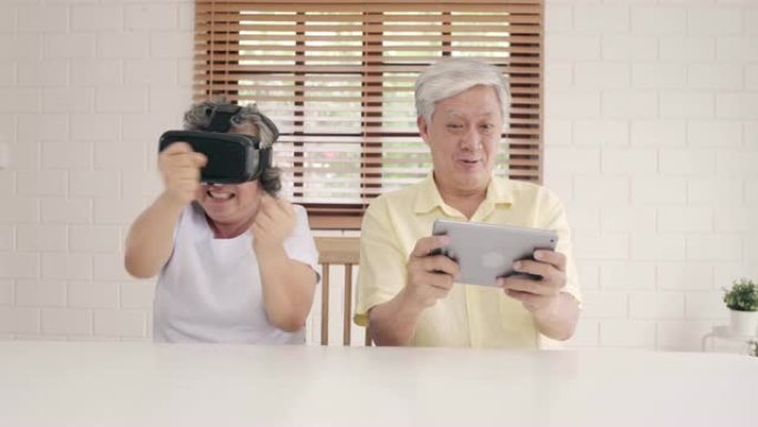 亚洲老年夫妇使用平板电脑和虚拟现实模拟器在客厅玩游戏，夫妇在一起躺在家里的桌子上感到快乐。生活方式高