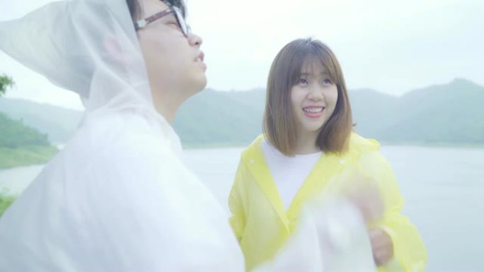 年轻的亚洲甜蜜夫妇穿着雨衣站在湖边，享受浪漫的雨水时光，感到快乐。生活方式夫妇在雨天享受和放松。