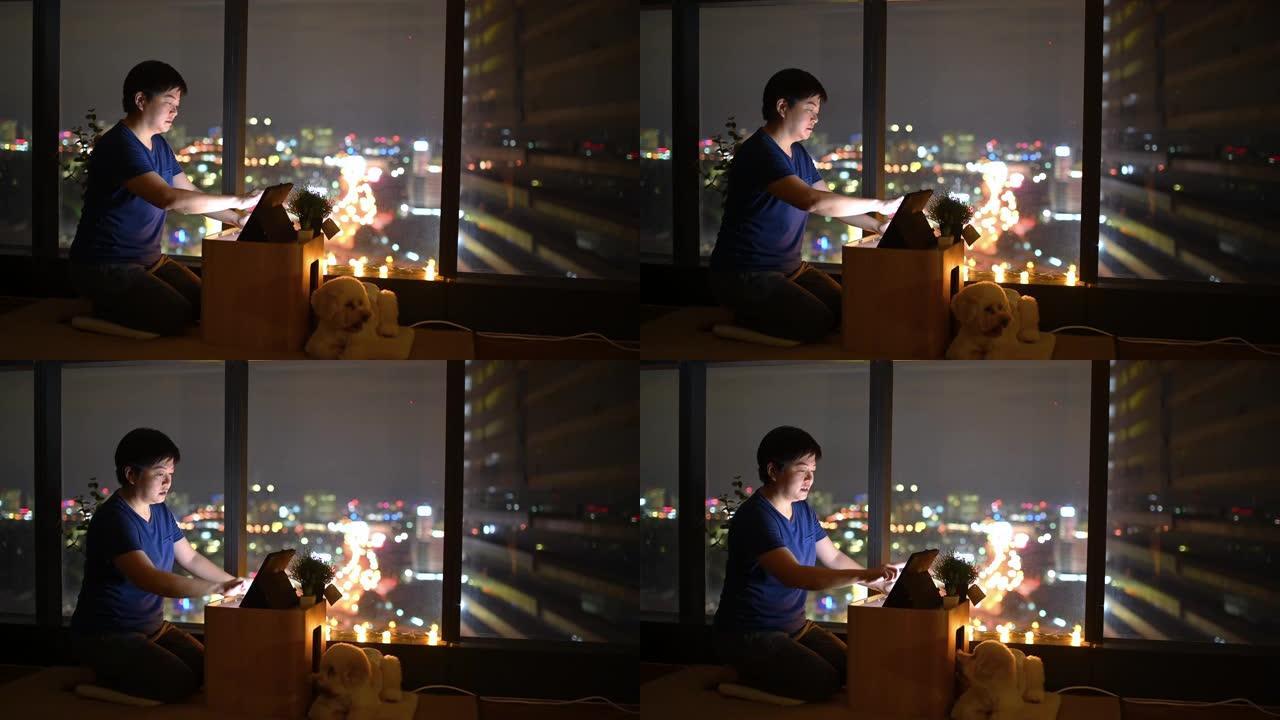 一个亚洲中国人晚上在窗户附近的公寓里玩数字平板电脑在线游戏，可以看到城市景观，玩具狮子狗宠物狗