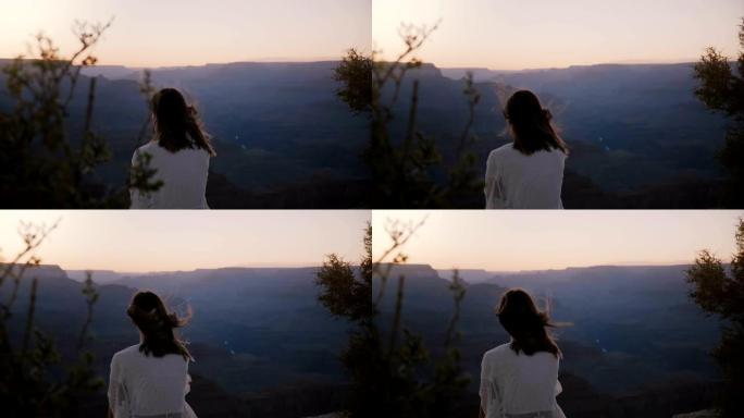 慢动作后视图快乐兴奋的年轻女子，头发在风中飘扬，坐在大峡谷上空史诗般的日落中。