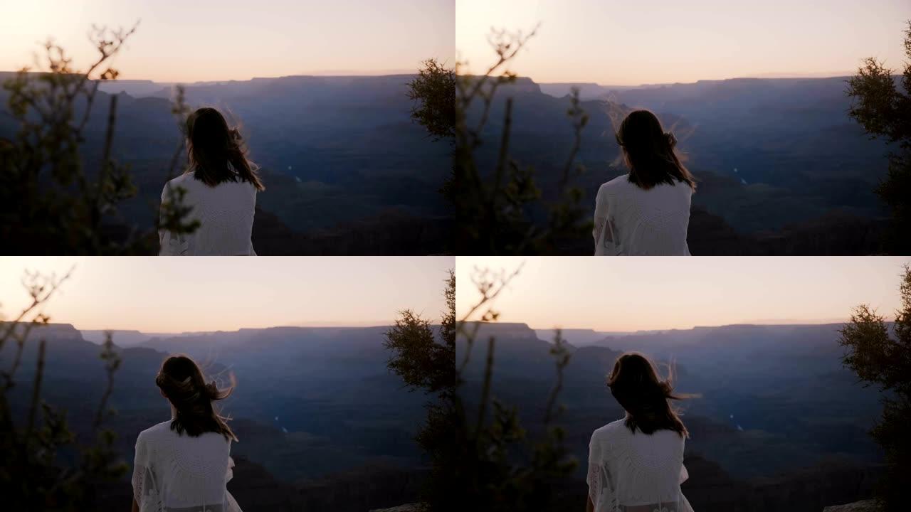 慢动作后视图快乐兴奋的年轻女子，头发在风中飘扬，坐在大峡谷上空史诗般的日落中。