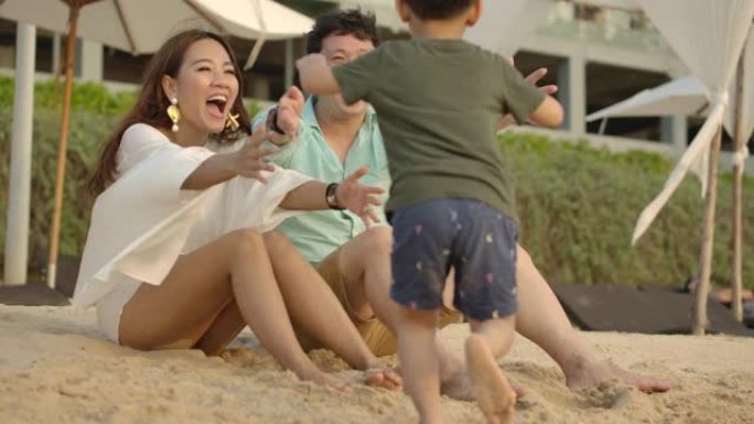 暑假可爱的亚洲家庭。一个小孩在海滩上跑向父母的慢动作镜头。然后父亲抱着他，拥抱，母亲亲吻亲爱的孩子。