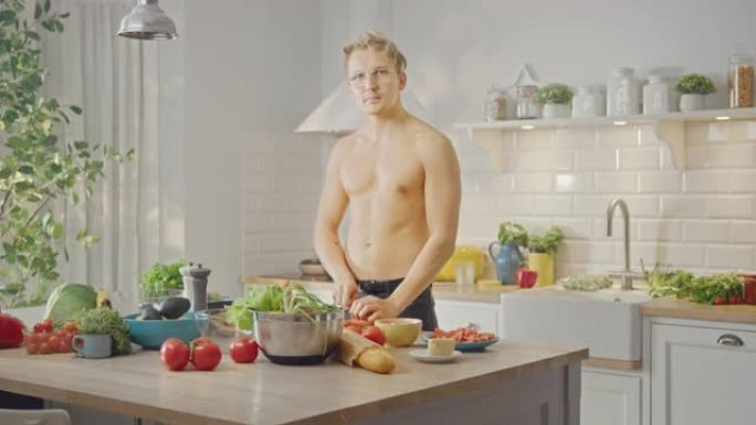 赤膊帅哥用锋利的菜刀切胡萝卜。在现代厨房里准备健康的有机沙拉餐。半裸减肥男对着镜头眨眼。