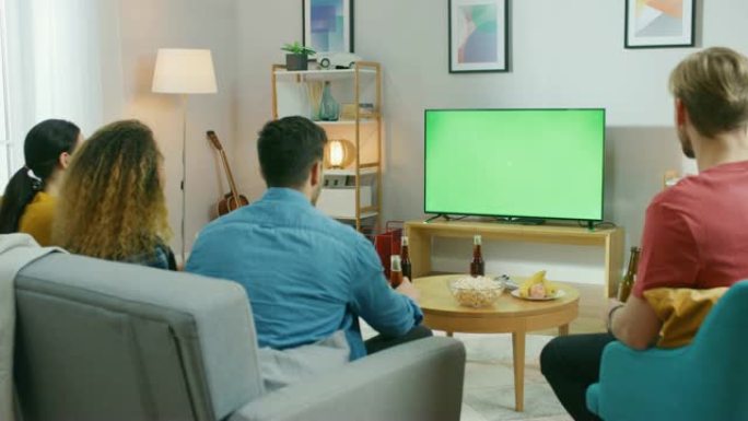 四个朋友坐在家里的沙发上，一边吃零食和喝饮料，一边看绿色色度键屏幕电视。年轻人在家里玩得开心。
