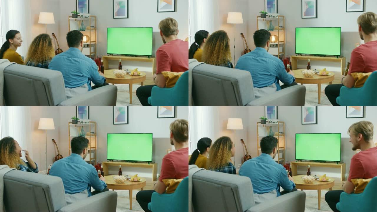 四个朋友坐在家里的沙发上，一边吃零食和喝饮料，一边看绿色色度键屏幕电视。年轻人在家里玩得开心。