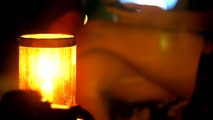 近距离拍摄美丽的蜡烛在晚上休息室酒吧的桌子上，女人的腿交叉使用智能手机在背景。