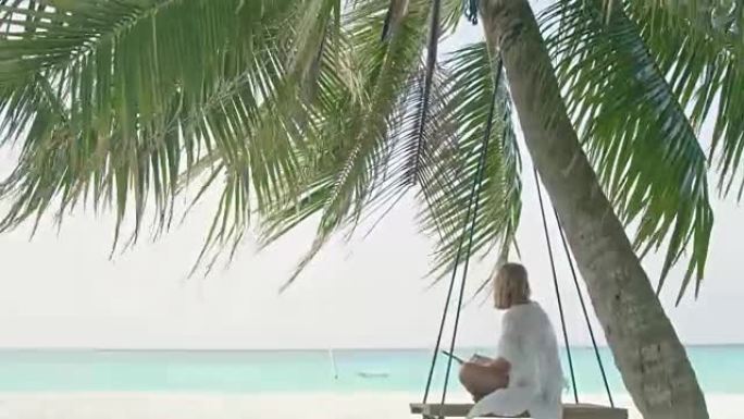 女士在马尔代夫热带海洋海滩田园诗般的棕榈树秋千上使用数字平板电脑