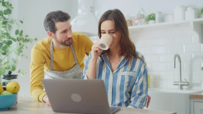 美丽的年轻夫妇在家里的厨房里使用笔记本电脑。穿着睡衣的女性。穿着围裙的男人鼓励女友选择购物。女性坐着
