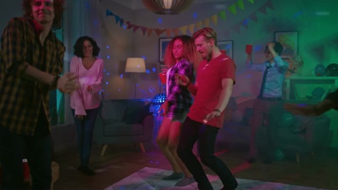 在大学之家聚会上: 性感的夫妇与周围有趣的朋友一起跳舞。男孩和女孩在客厅里泡吧。迪斯科霓虹灯频闪灯照