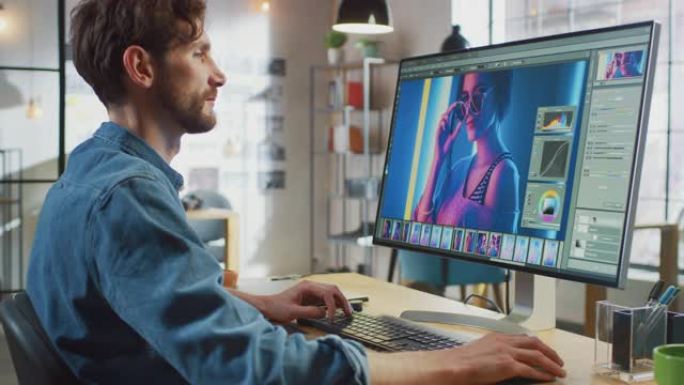 穿着牛仔裤衬衫的男性数字编辑器在他的个人电脑上大显示屏的图像编辑软件中工作。他在凉爽的办公室阁楼工作