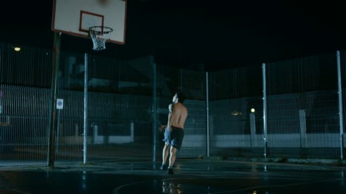 运动年轻的男子篮球运动员运球并在围栏的街球场上扔球。庆祝打进两分球。居民区雨后的夜间录像。