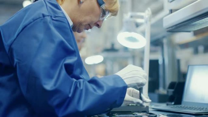 穿着蓝色工作服和防护眼镜的女电子工厂工人正在用螺丝刀组装智能手机。高科技工厂设施，后台有更多员工。