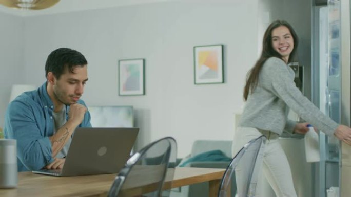 美丽的年轻夫妇在家里的厨房里聊天。男人正在用笔记本电脑工作，女孩从冰箱里拿出一个瓶子，把饮料倒入杯子