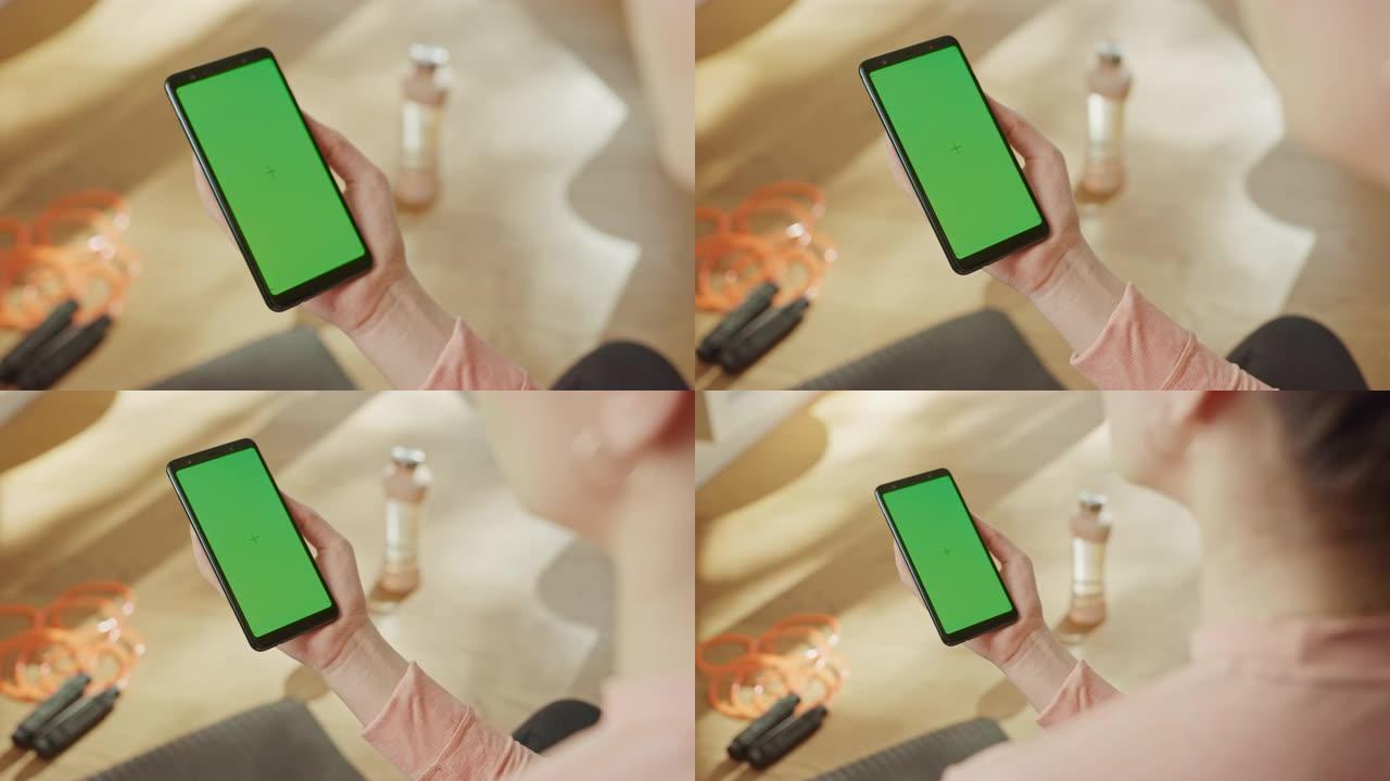 健身概念: 女人手持色度键绿屏智能手机。背景健身垫，跳绳和设备。特写视点视点