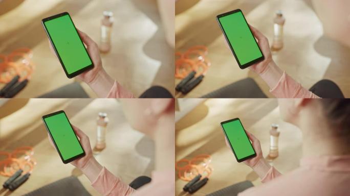 健身概念: 女人手持色度键绿屏智能手机。背景健身垫，跳绳和设备。特写视点视点