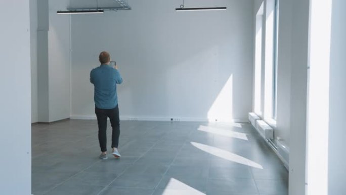 穿着牛仔裤衬衫的年轻时髦男子站在空荡荡的办公室里，用平板电脑上的增强现实软件对其进行映射。阳光透过大