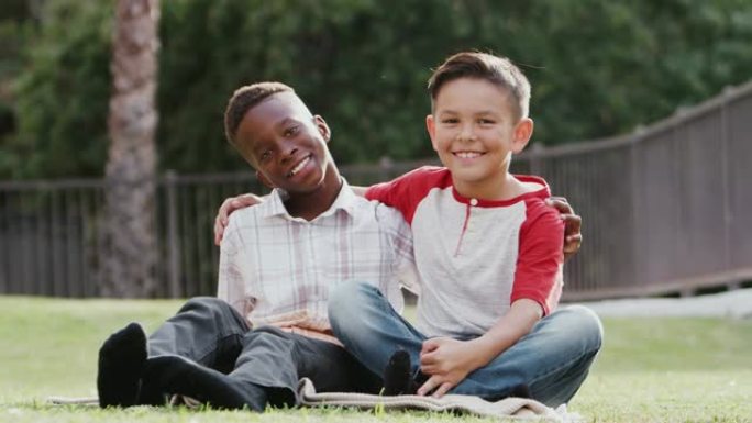 两个青春期前的男性朋友坐在草地上，双臂环抱，对着镜头微笑