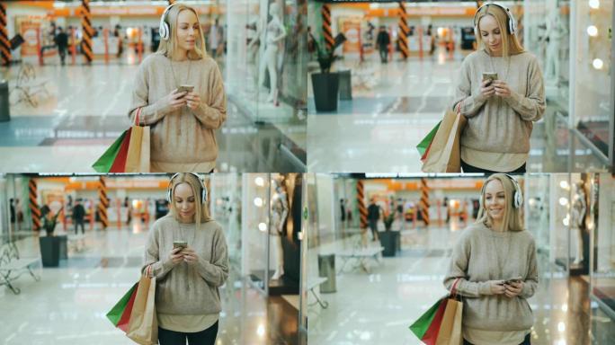 留着金色长发的快乐年轻女子正在戴着耳机听音乐，一边拿着纸袋在购物中心散步，一边使用智能手机。