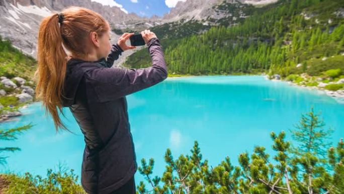 女人在山上徒步旅行，用智能手机拍摄美丽的蓝色湖泊的壮丽景色