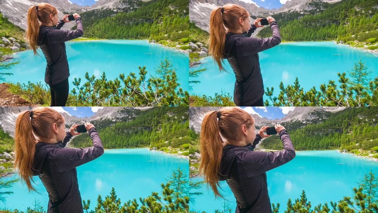女人在山上徒步旅行，用智能手机拍摄美丽的蓝色湖泊的壮丽景色