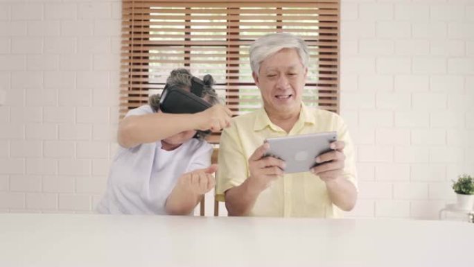 亚洲老年夫妇使用平板电脑和虚拟现实模拟器在客厅玩游戏，夫妇在一起躺在家里的桌子上感到快乐。生活方式高