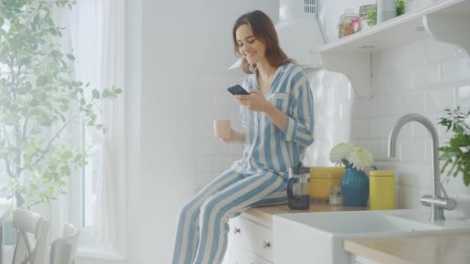 美丽的女性坐在橱柜上，在厨房里使用智能手机，同时喝着一杯刚煮好的咖啡。穿着睡衣、生活方式健康的女孩早