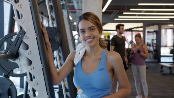 健身房里瘦瘦的年轻女子对着镜头兴高采烈地拿着一个水瓶靠在健身器上
