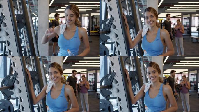 健身房里瘦瘦的年轻女子对着镜头兴高采烈地拿着一个水瓶靠在健身器上