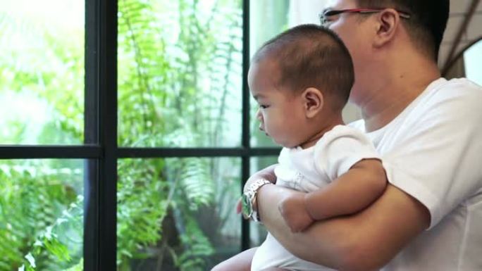 4k慢动作镜头，亚洲男婴凝视着窗户玻璃，而他的父亲则在现代阁楼，家庭和生活方式的概念