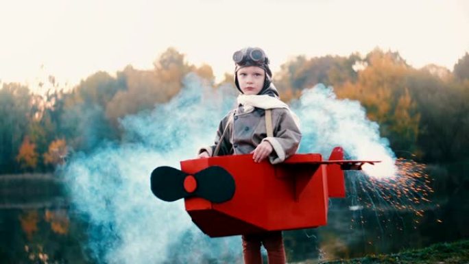 快乐的小飞行员男孩站在日落湖，穿着彩色烟雾的纸板飞机服装玩飞行员慢动作。