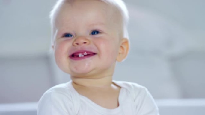 一个婴儿，一个有着蓝色大眼睛和浅色头发的男孩，坐在雪白的毯子上微笑，看着白色背景上的母亲。