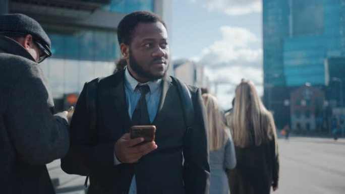 一个穿着西装的非洲裔美国商人的肖像，站在街上，有行人。他在用智能手机。他看起来很成功。其他人去上班。