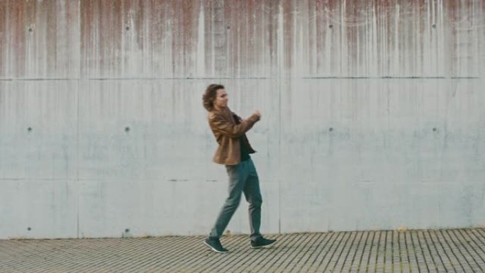 开朗快乐的长发小伙子在城市混凝土墙旁边的街道上散步并积极跳舞。他穿着一件棕色皮夹克。晴天。
