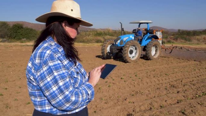 女农民使用数字平板电脑并监视拖拉机在大型蔬菜农场的农作物上喷洒农药
