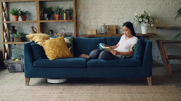 美丽的亚洲女孩正在阅读客厅沙发上的书，而机器人吸尘器则是干洗地板和地毯。技术和人的概念。