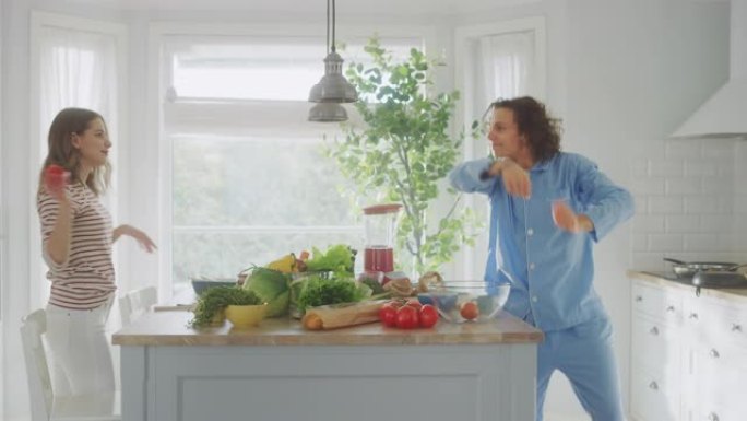 穿着蓝色睡衣的英俊年轻人和漂亮的女孩在厨房里跳舞。幸福的夫妻正在互相摆弄绿色蔬菜。晴天在家男女。
