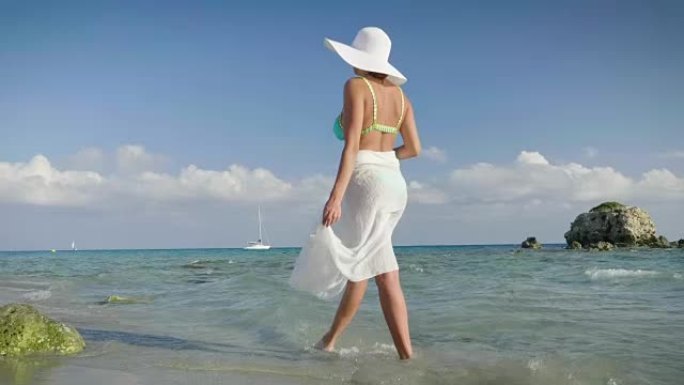 一个美丽的年轻女孩在海边漫步，穿着一顶草帽的泳衣，一条白色的方巾，金色的沙子，山峦和大海的背景。