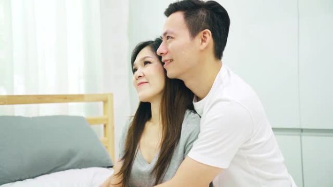 4K UHD: 年轻的亚洲夫妇坐在床上，看着窗外思考。沉思概念